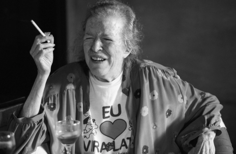 Escritora Hilda Hilst. Foto em preto e branco. Ela é uma mulher velha, de cabelos desgrenhados, usa um roupão florido aberto e, por baixo, uma camiseta estampada "Eu amo vira-latas". Ela está sorridente, sentada de jeito confortável, segurando um cigarro com a mão direita.