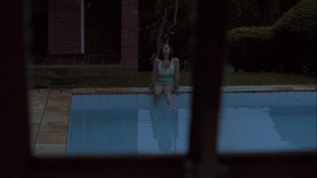 Cena do filme Irene. Através de uma janela, vemos a mulher sentada na beira da piscina com um maiô verde. Ela olha para cima com um sorriso e balança os pés na água.