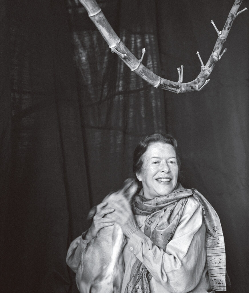Retrato em preto e branco de Hilda Hilst aos 60 anos. Ela sorri para a câmera e afaga um cachorro, que parece querer lamber o seu rosto.