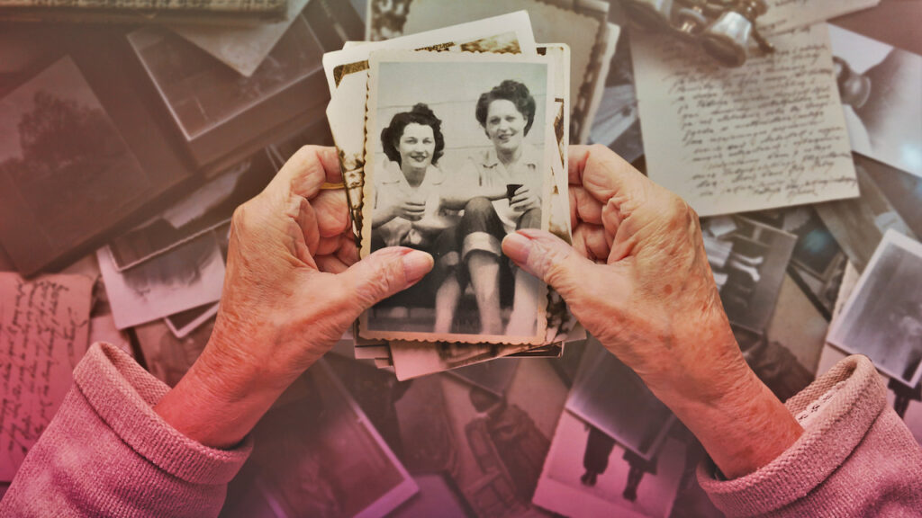 A fotografia mostra duas mãos brancas e velhas segurando um bolo de fotos antigas, em preto e branco. A foto que vemos, junto com a dona das mãos, mostra duas mulheres sentadas lado a lado, olhando para a câmera e sorrindo.